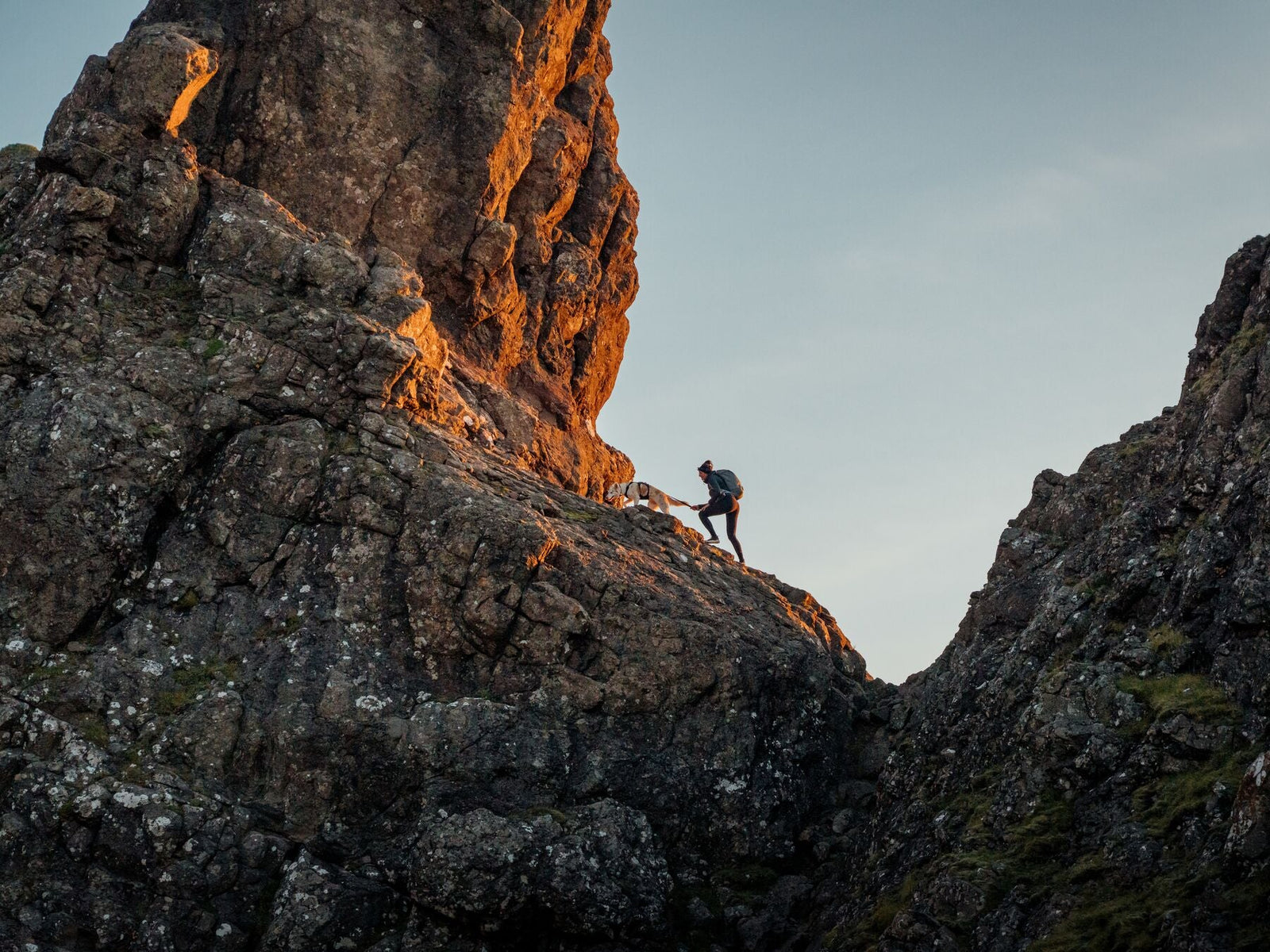 A hiker and their dog scrambling together over dramatic rocky terrain at golden hour, wearing Ruffwear adventure gear