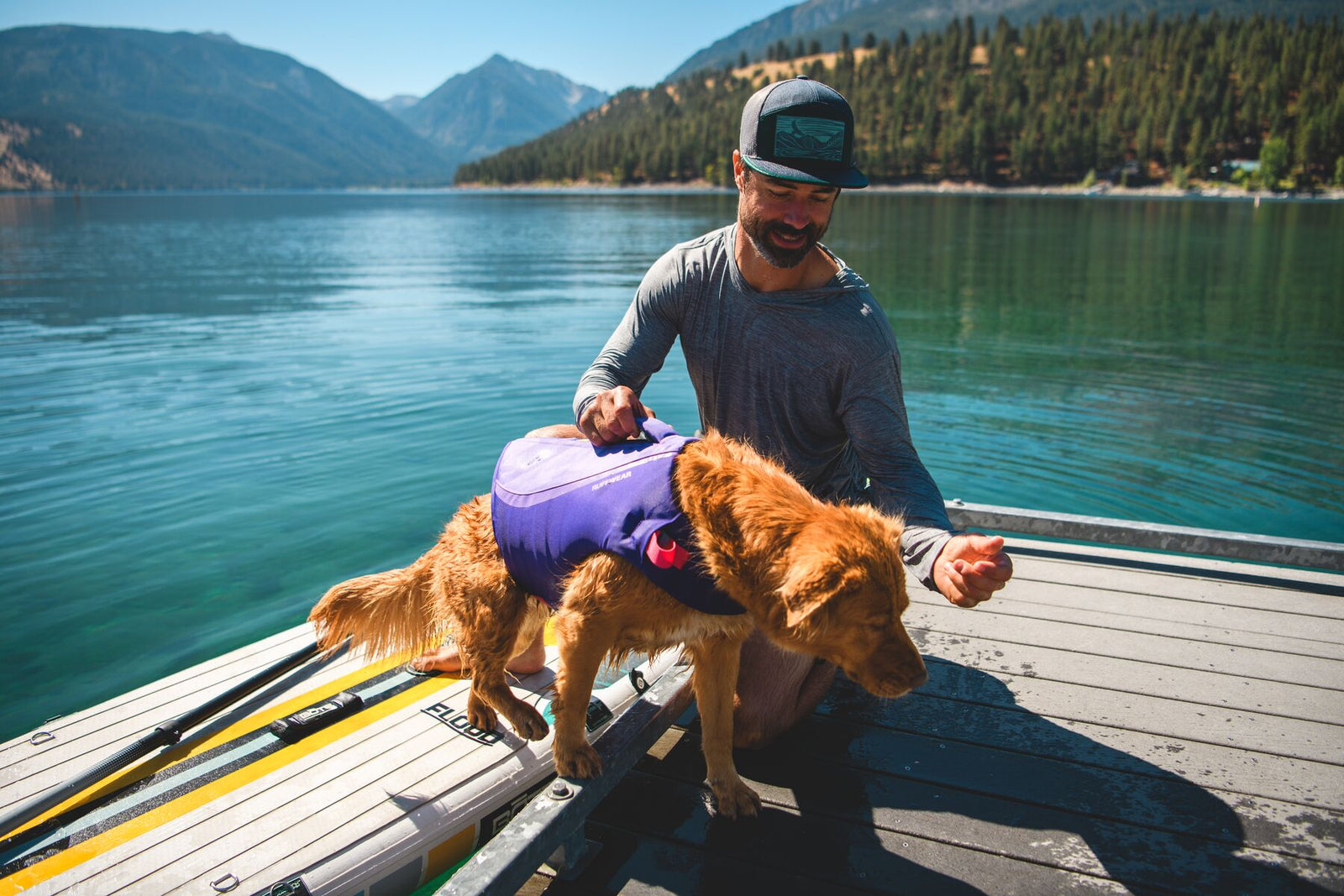 Person lifting a golden retriever wearing the Ruffwear Float Coat in Purple Lily by the handle, on a dock by a mountain lake.