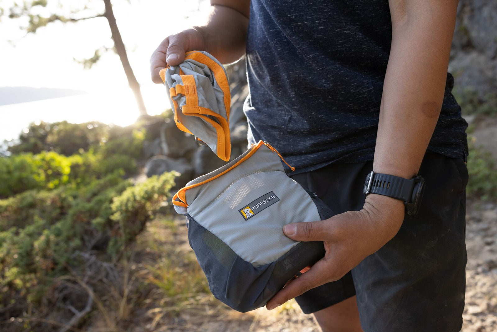 Hiker unpacking the Ruffwear BackTrak Evac Kit from its compact stow pocket on a rocky trail at golden hour, lake and pine trees visible behind, showing how small and lightweight the kit packs down