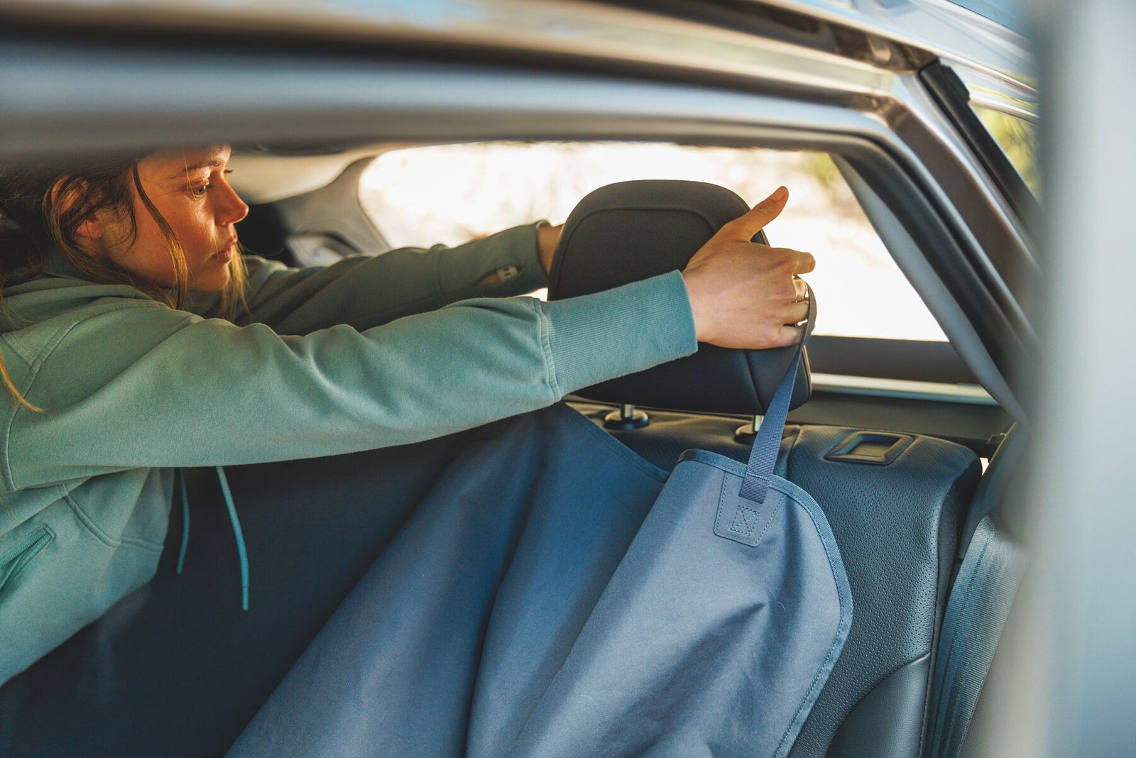 Woman in a sage green jacket fitting the Ruffwear Dirtbag Seat Cover over the back seat headrest of a car, cover laid across the back seat in bench configuration, warm golden light through the window