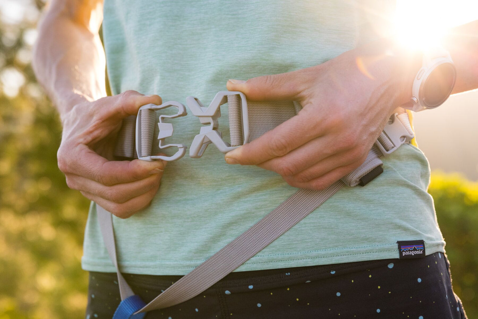 Close-up of hands fastening the ITW Nexus buckle on the Ruffwear Trail Runner Belt against a runner's waist, warm golden backlight through trees, blue Trail Runner Leash visible at the bottom of frame