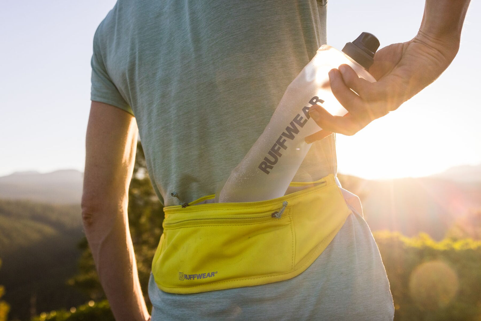 Runner pulling a Ruffwear soft flask from the zippered pocket of the Ruffwear Trail Runner Belt in Lichen Green, worn at the waist against a golden sunset over open hills, showing the stretch pocket capacity in use
