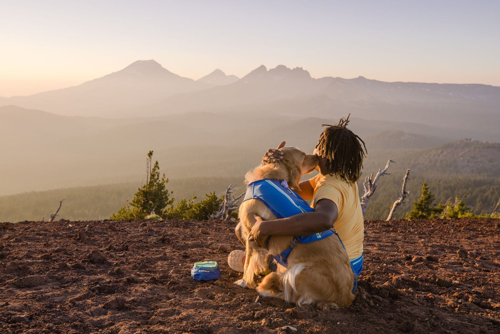 Person and dog sitting together on a mountain summit at dusk, the dog wearing the Ruffwear Trail Runner Running Vest in Blue Pool, misty mountain range stretching into the distance behind them