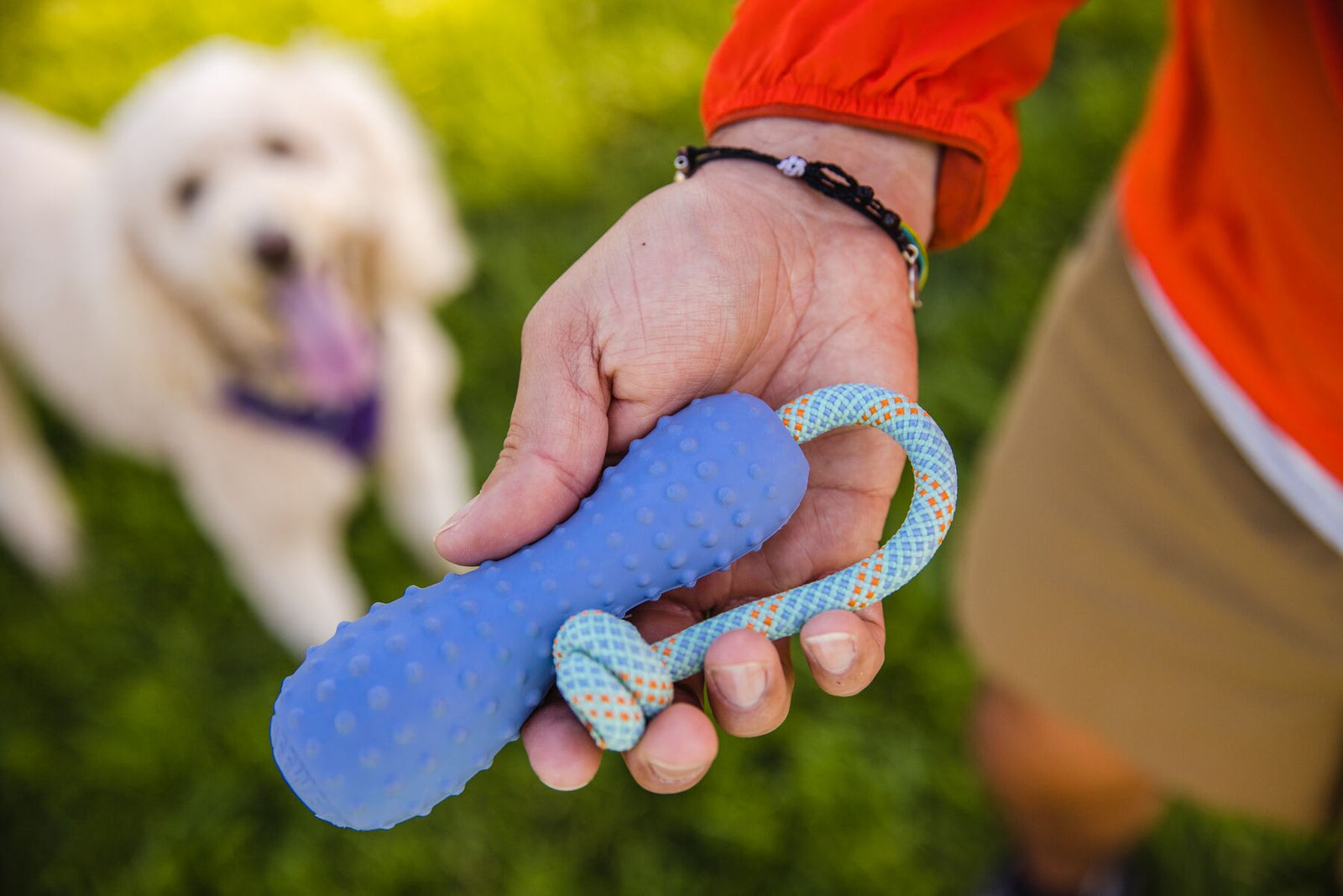 Person holding the Ruffwear Gourdo dog toy in Heliotrope Purple by the kernmantle rope handle outdoors, with a happy white dog visible in the background