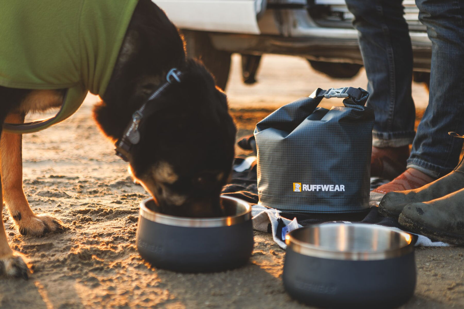 Black dog in a green Ruffwear coat eating from a Ruffwear Basecamp Bowl on a sandy beach at golden hour, with the Ruffwear Kibble Kaddie in Basalt Gray sitting upright alongside and a second Basecamp Bowl in the foreground, campervan visible behind them