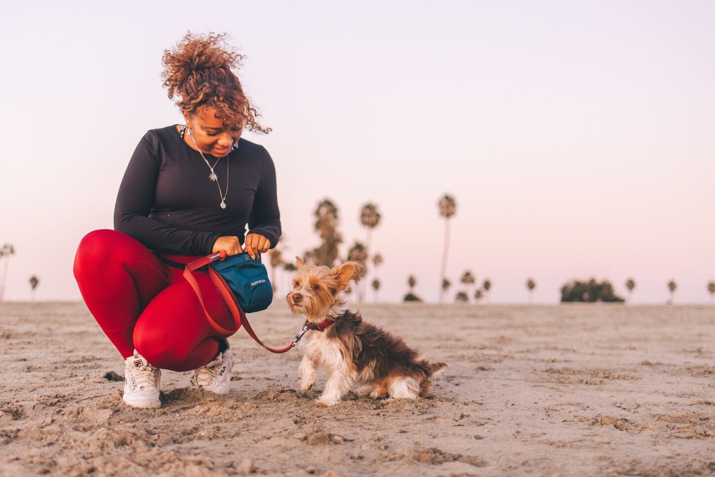 Woman crouching on a sandy beach at dusk with a small Yorkshire Terrier on a red lead, opening the Ruffwear Pack Out Bag in blue to stow a dog waste pick-up bag, palm trees visible in the background