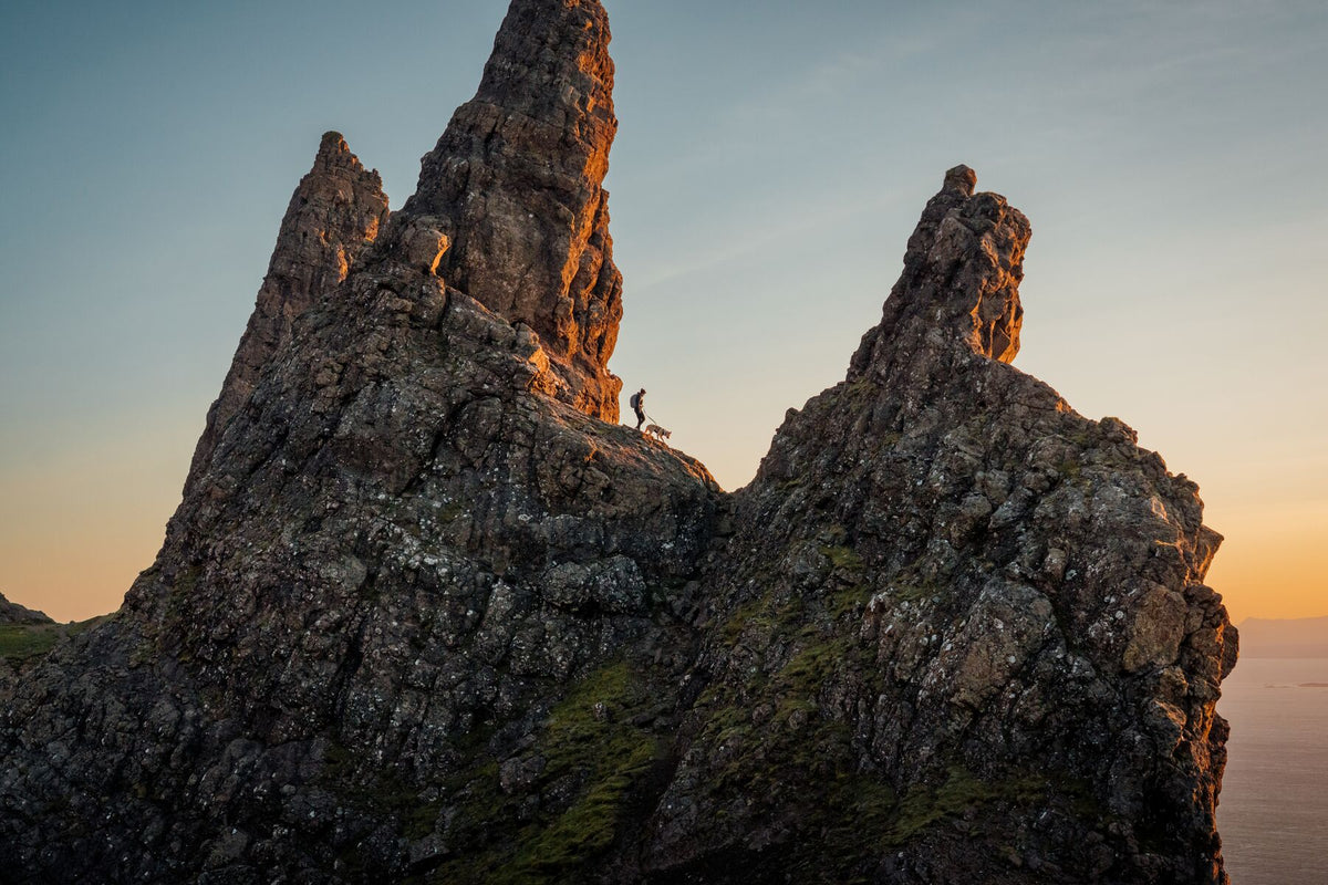 Person and dog picking their way along a narrow rocky ridge at sunset, towering basalt rock formations rising around them, warm golden light catching the stone - the Ruffwear Ridgeline harness built for exactly this