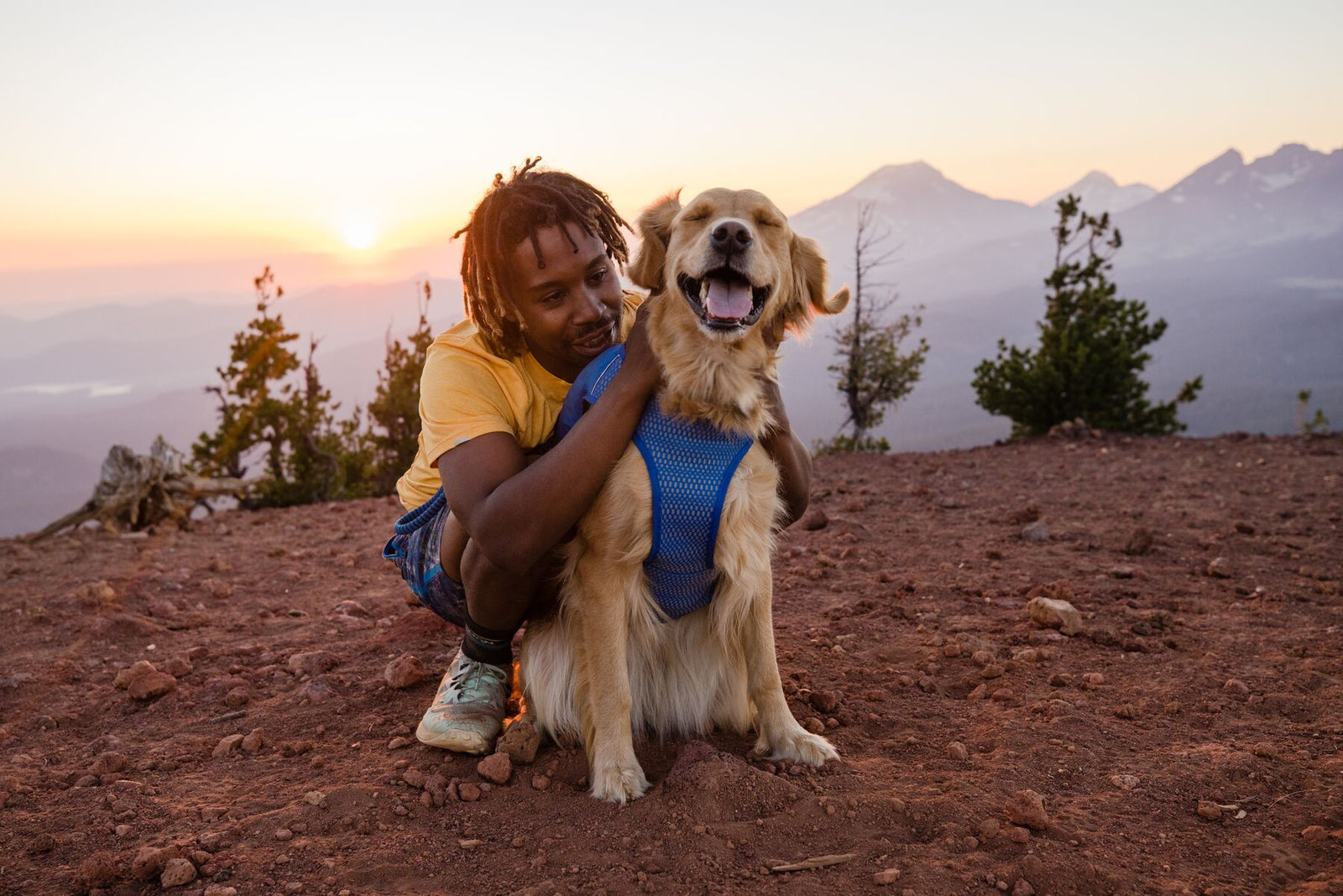 Man crouching beside a happy golden retriever wearing the Ruffwear Trail Runner Running Vest in Blue Pool on a mountain summit at sunset, peaks and open sky stretching behind them