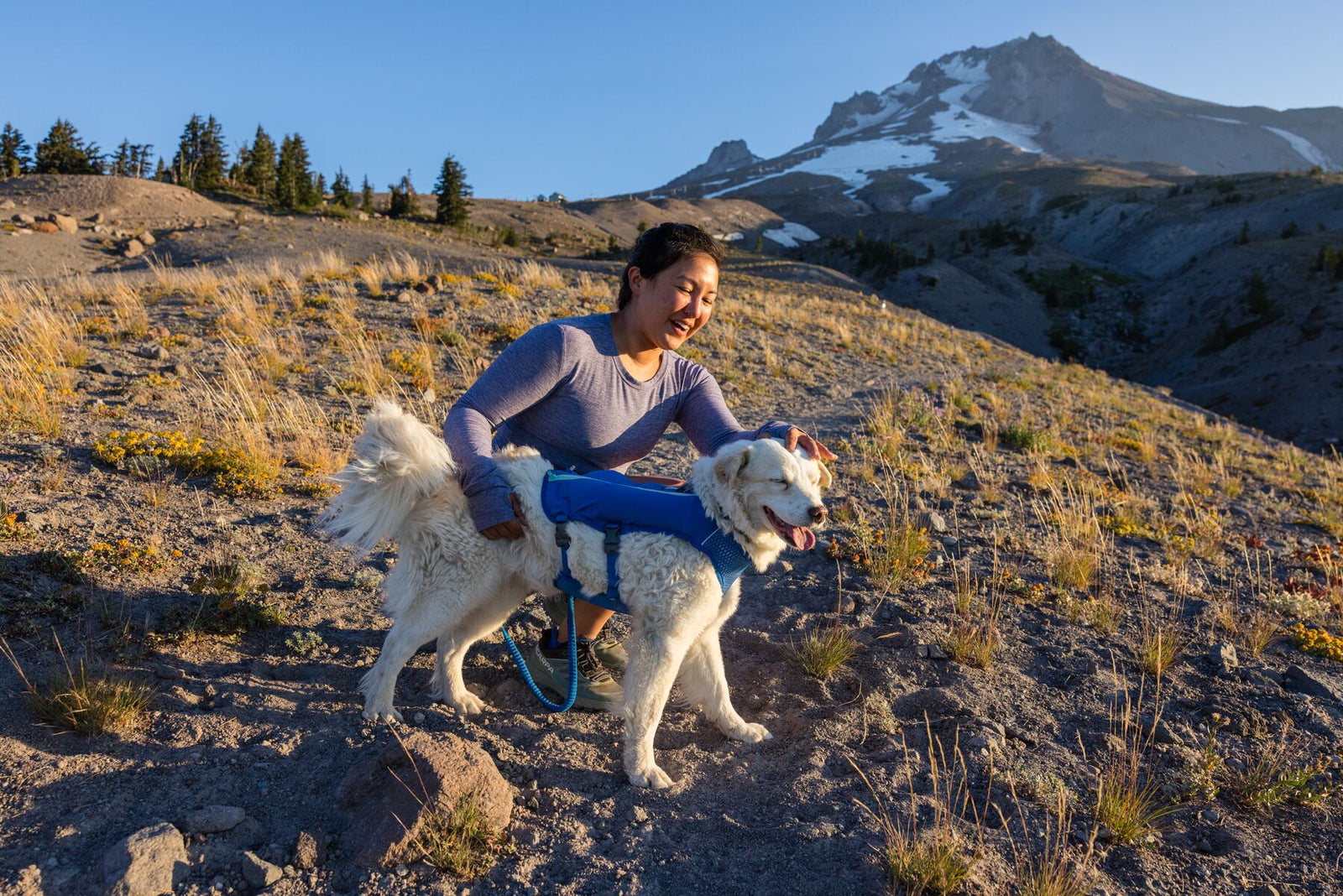 Woman crouching beside a large white fluffy dog wearing the Ruffwear Trail Runner Running Vest in Blue Pool on an open mountain trail, snow-capped peak rising behind them under a clear blue sky