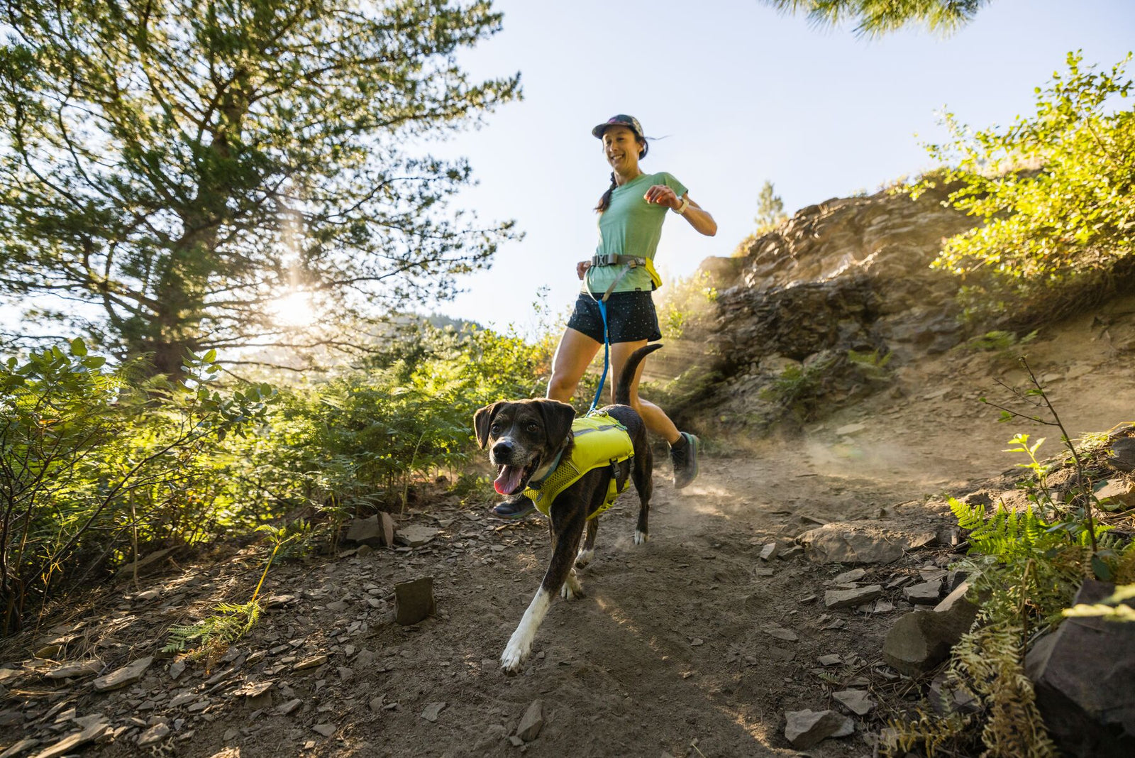 Woman trail running uphill on a rocky woodland path with a dark dog wearing the Ruffwear Trail Runner Running Vest in Lichen Green, sun flaring through the trees behind them