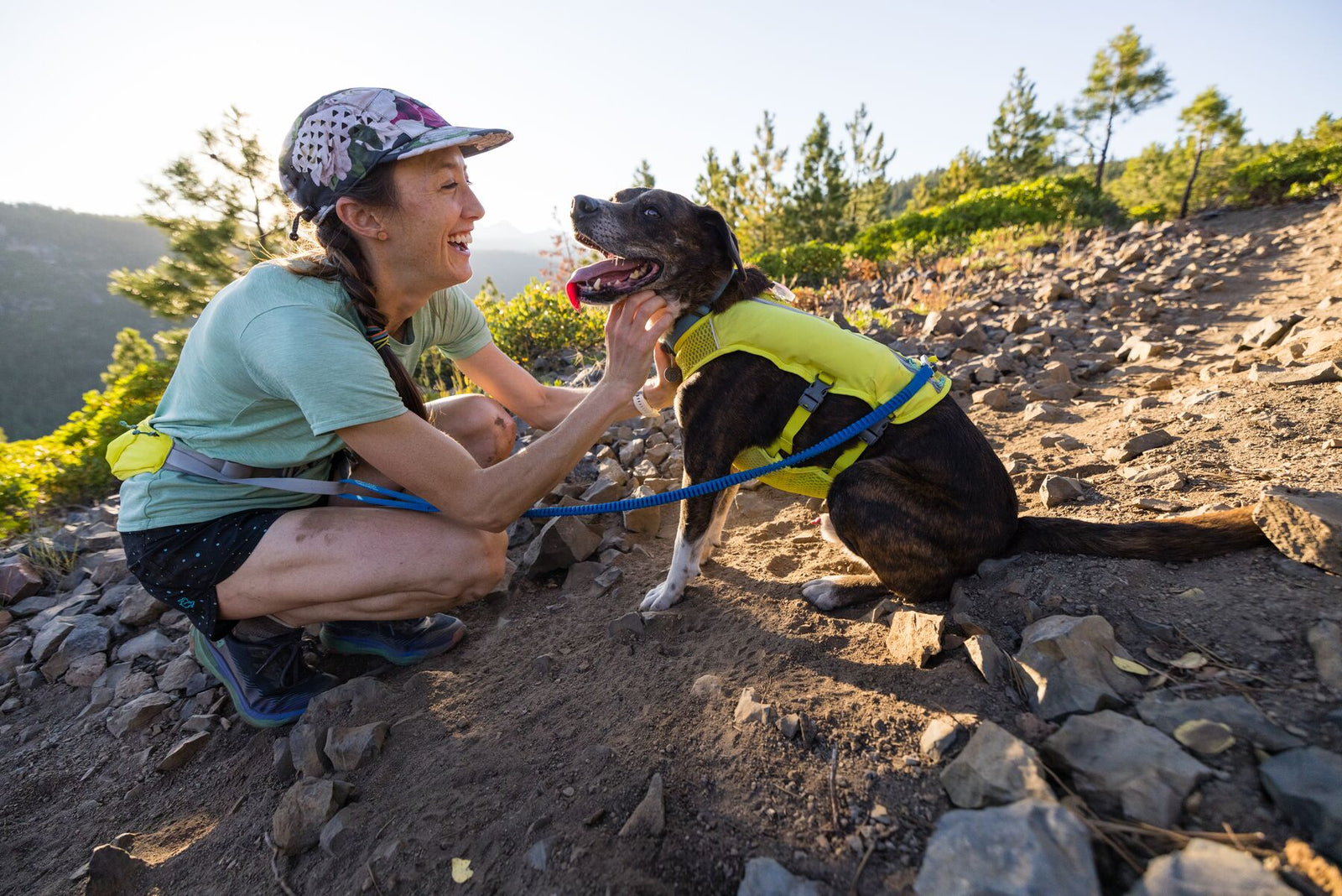 Woman crouching on a rocky trail laughing with a dark brindle dog wearing the Ruffwear Trail Runner Running Vest in Lichen Green, wooded hillside and open valley stretching behind them in the evening light